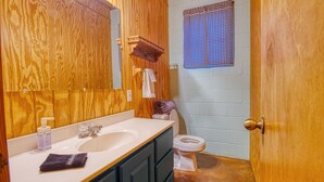 Un baño con paredes de madera, un lavabo blanco, un espejo, un perchero y una ventana con cortinas cuadriculadas.