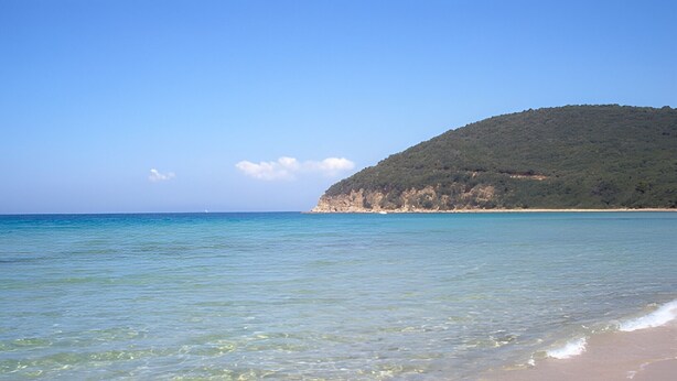 A beach with clear water, a sandy shore, and a hill in the background.
