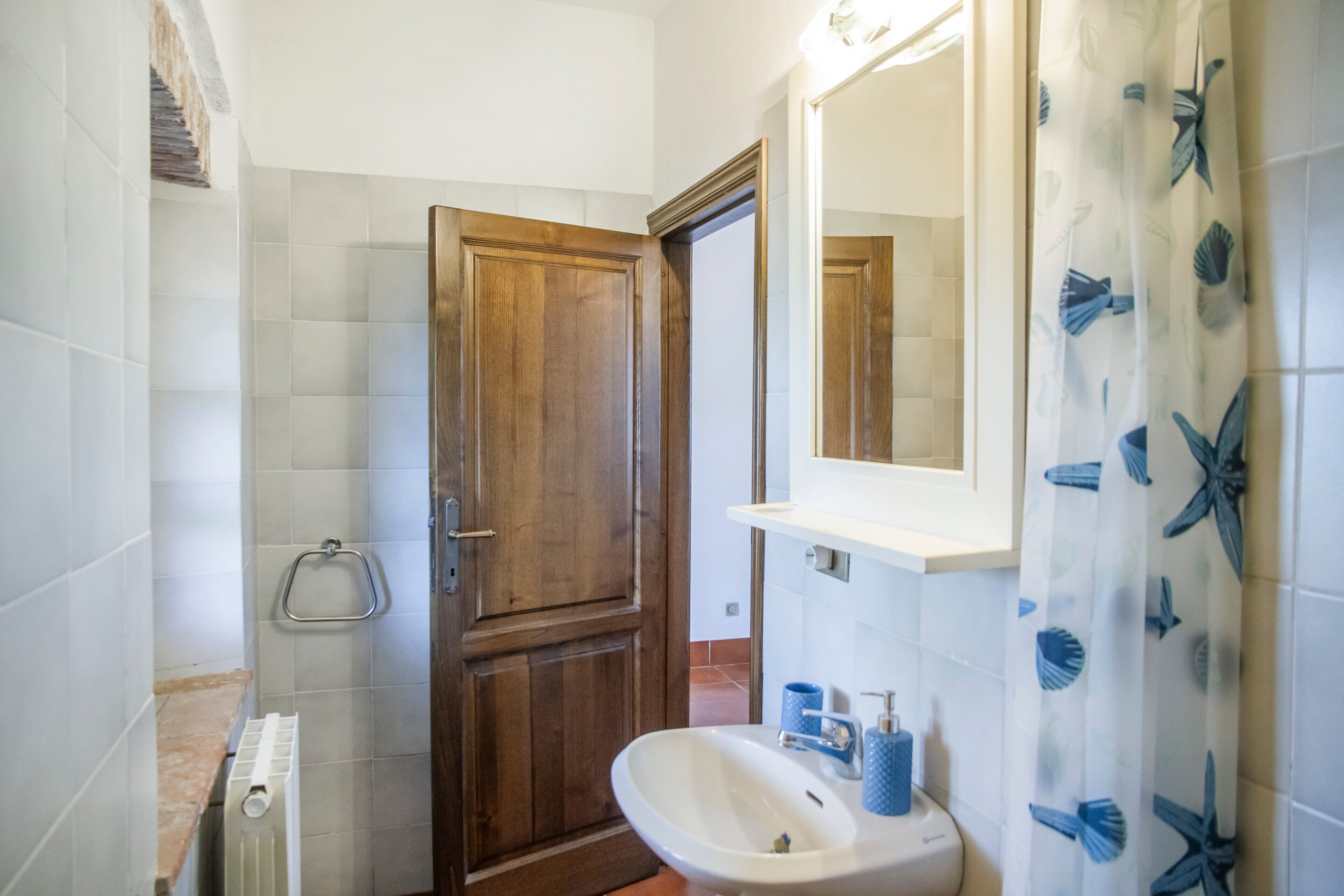 Un baño con puerta de madera, lavamanos blanco, espejo y cortina de ducha con diseño de hojas.