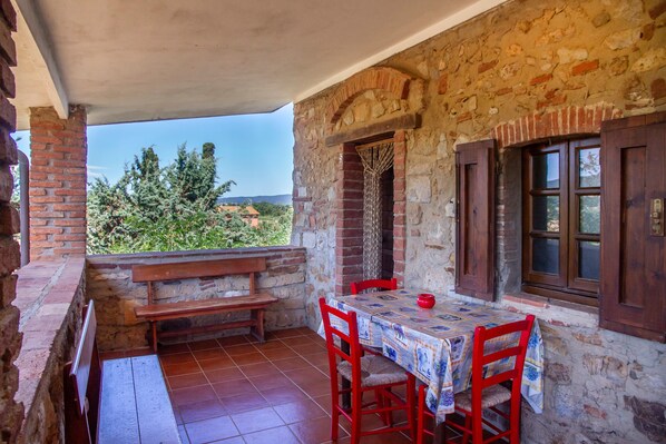 A stone-walled room with a wooden table and chairs, a bench, and a view of greenery.