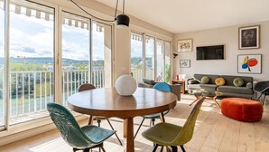 A spacious living room with a round wooden table, colorful chairs, a red ottoman, and a grey sofa. Large windows offer a view of a river and distant buildings.
