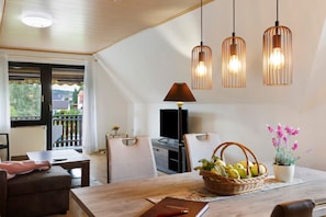 Un coin repas comprenant une table, des chaises, un panier de fruits, une plante en pot et des luminaires suspendus.