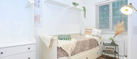 Une chambre à coucher comprenant un lit, une commode, un radiateur, une fenêtre avec des stores et une petite table avec une lampe.