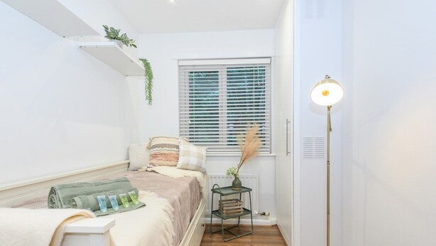 Un dormitorio con cama, mesita de noche, lámpara, ventana y radiador.