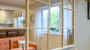 Una sala de estar acogedora con un sofá rosa, un comedor con sillas de madera, una cocina al fondo y una ventana con vistas a zonas verdes.