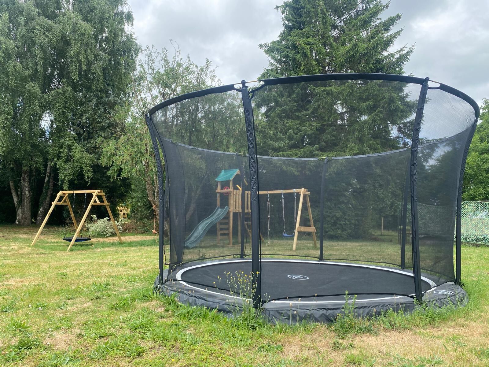 Un trampolín con red de protección rodeado de césped, con un conjunto de columpios de madera y un tobogán al fondo.