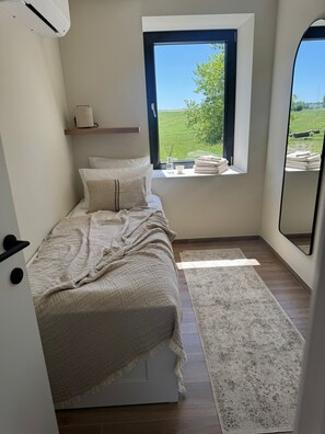 Een slaapkamer met een bed, een raam met uitzicht op groen, een nachtkastje met een lamp en een spiegel.