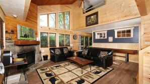Ein Holzblockhaus-Interieur mit Kamin, zwei Ledersofas, einem Couchtisch und einem großen Fenster mit Blick auf den Wald.
