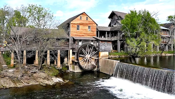 Ein historisches Holzgebäude mit einem großen Wasserrad und einer Holzbrücke über einem Fluss.