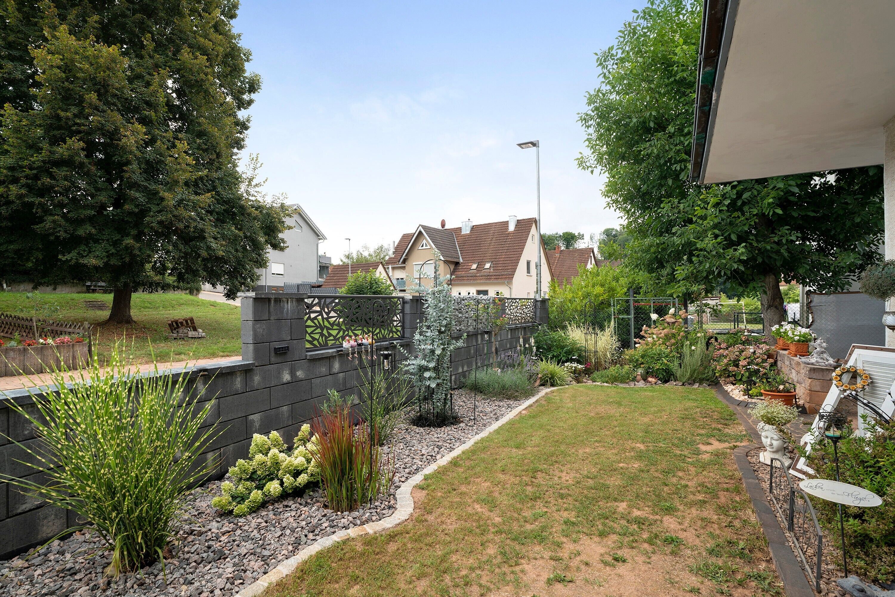 Ein Wohngarten mit Rasen, verschiedenen Pflanzen, einem Zaun und einem Haus im Hintergrund.