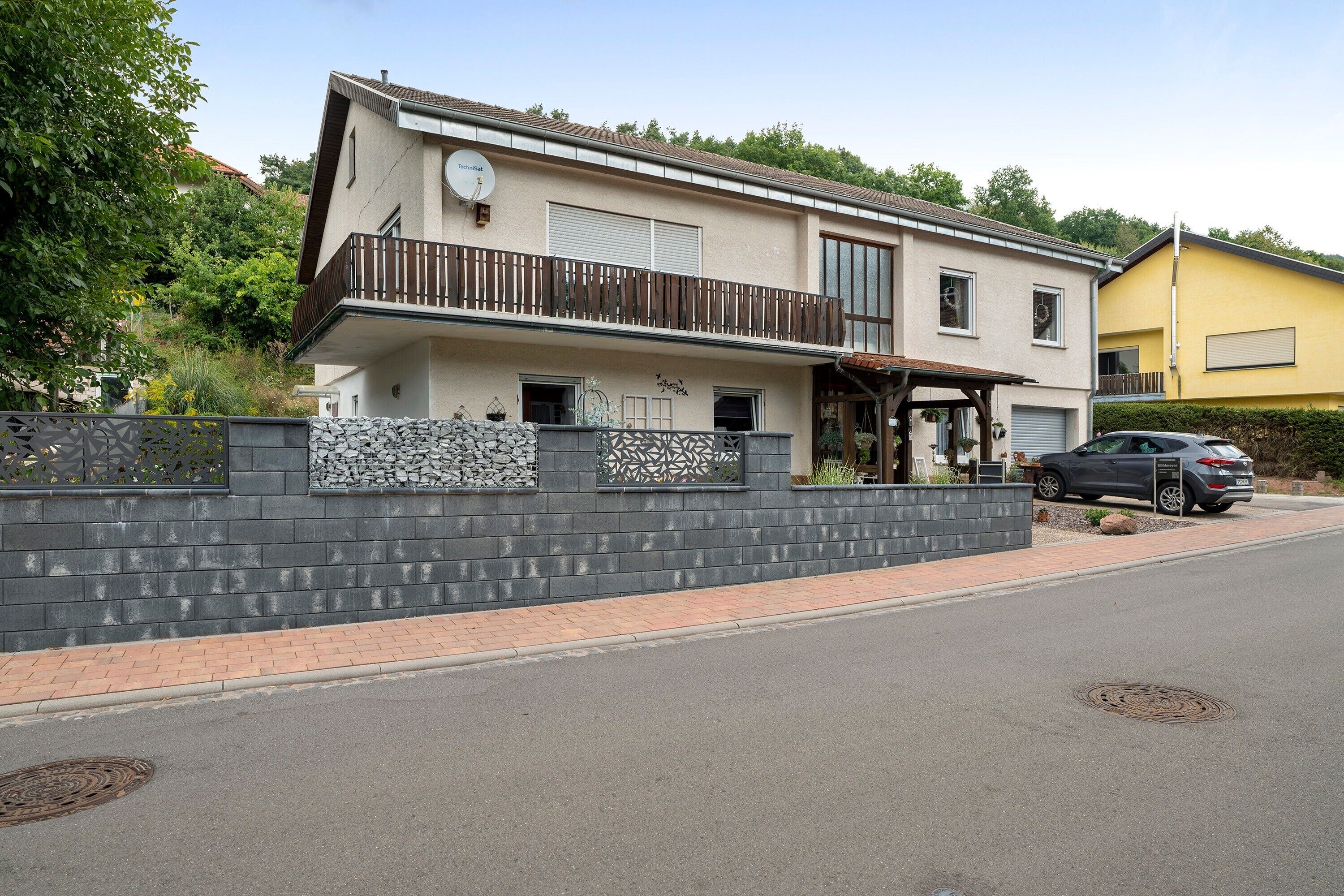 A two-story house with a balcony, a car parked in front, and a stone wall.