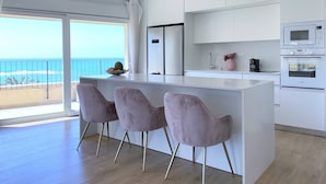 A modern kitchen with white cabinets, a large island with bar stools, a refrigerator, and an oven. A window offers a view of the beach. - 1604 Tel aviv (Tel Aviv)