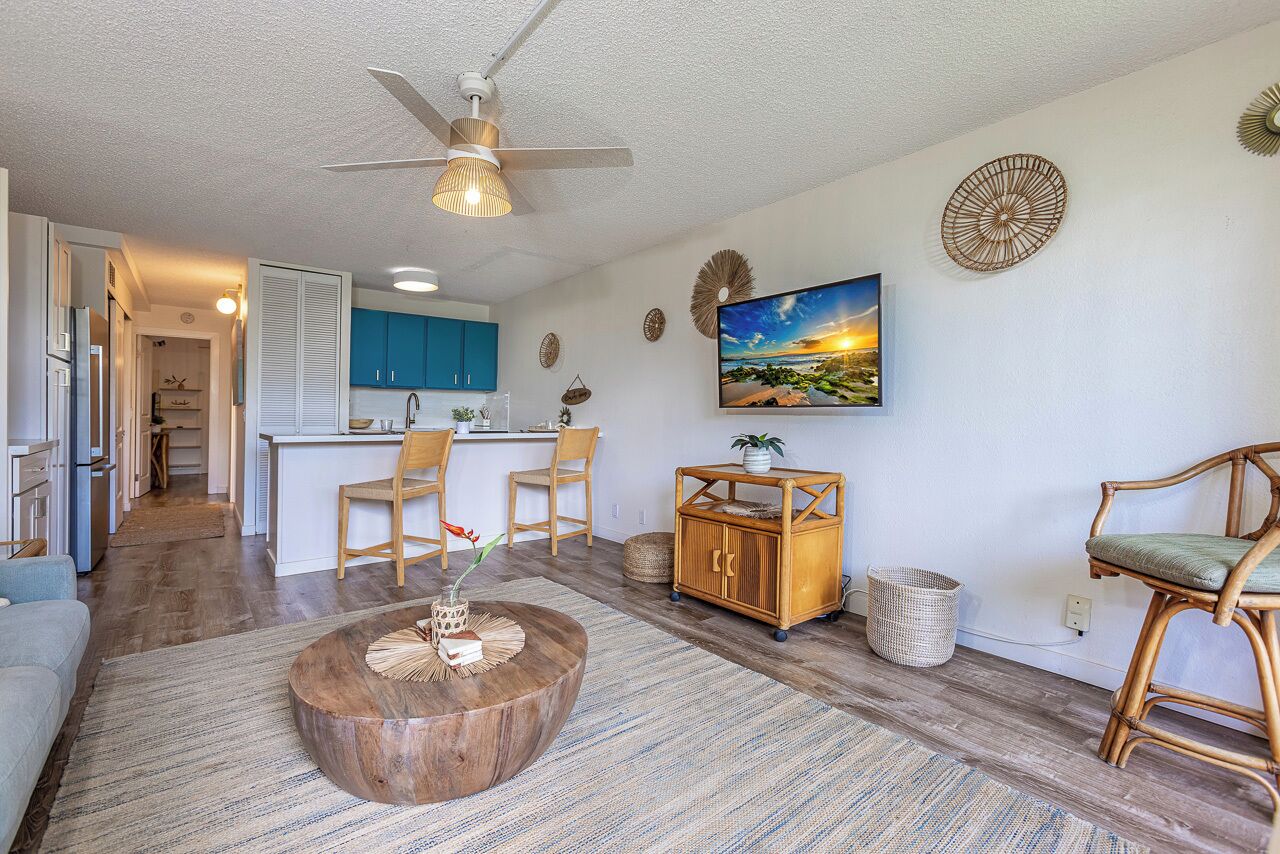 A modern living room with a wooden coffee table, a ceiling fan, and a kitchen area with blue cabinets and a dining table.