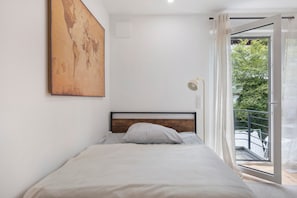 A bedroom with a bed, a wooden headboard, a large abstract painting, a window with a view of greenery, and a balcony.