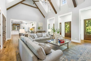 A spacious living room with a large sectional sofa, a glass coffee table, and a view of the kitchen and dining area through an open doorway.