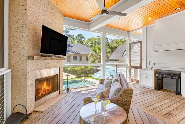 A modern outdoor living area with a fireplace, a television, a pool, and a grill.