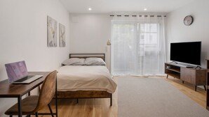 Een slaapkamer met een bed, een bureau met een laptop, een televisie, een stoel en een raam met gordijnen.
