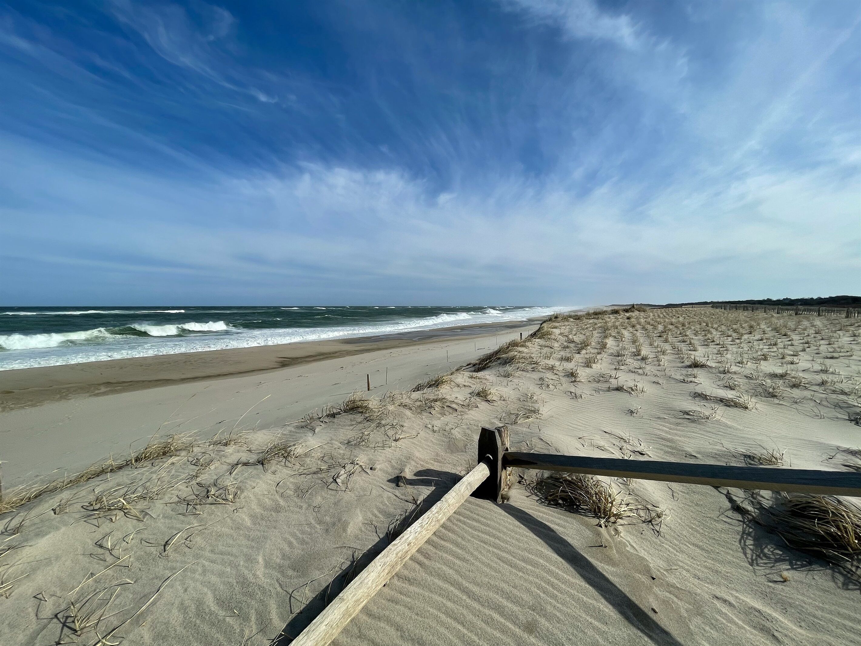 Une plage de sable où les vagues viennent se briser sur le rivage, une clôture en bois au premier plan et un ciel dégagé.