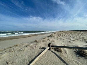 Una playa arenosa con olas rompiendo en la orilla, una valla de madera al frente y un cielo despejado.