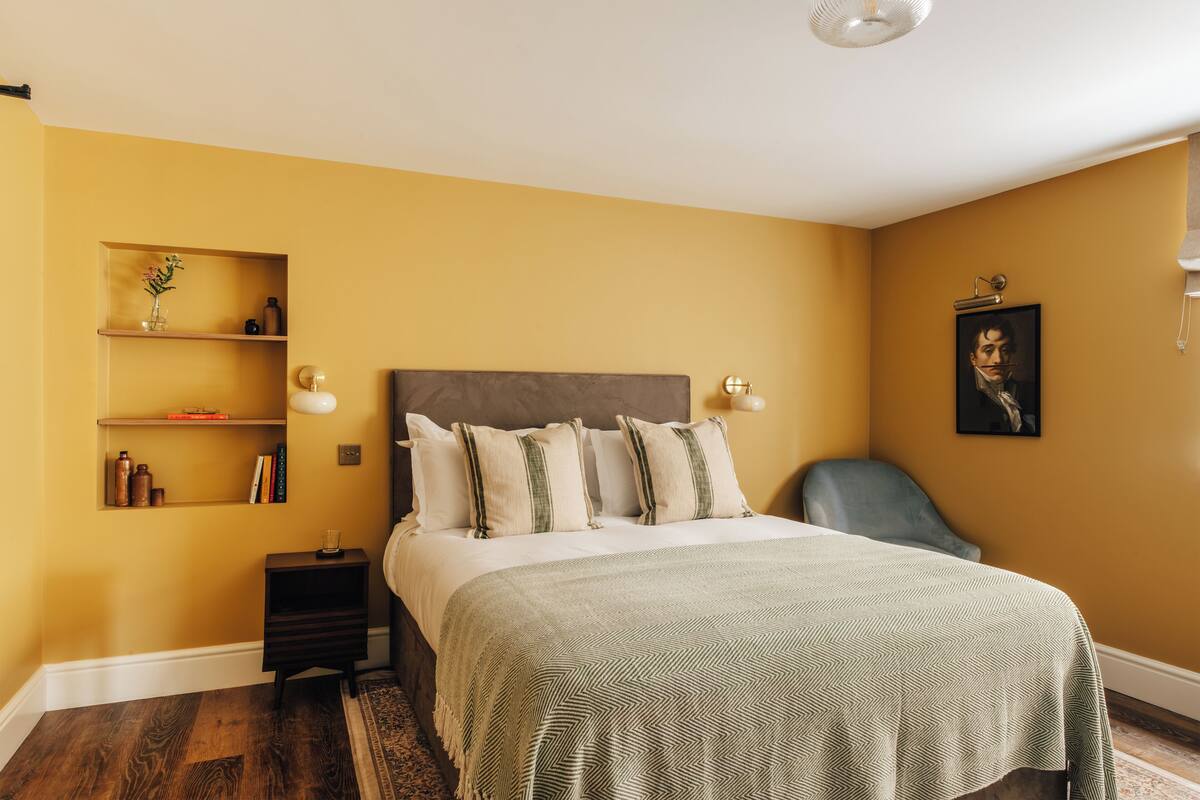 Une chambre à coucher comprenant un lit, des tables de chevet, une chaise, une étagère avec des livres et des objets décoratifs, et un tableau encadré accroché au mur.