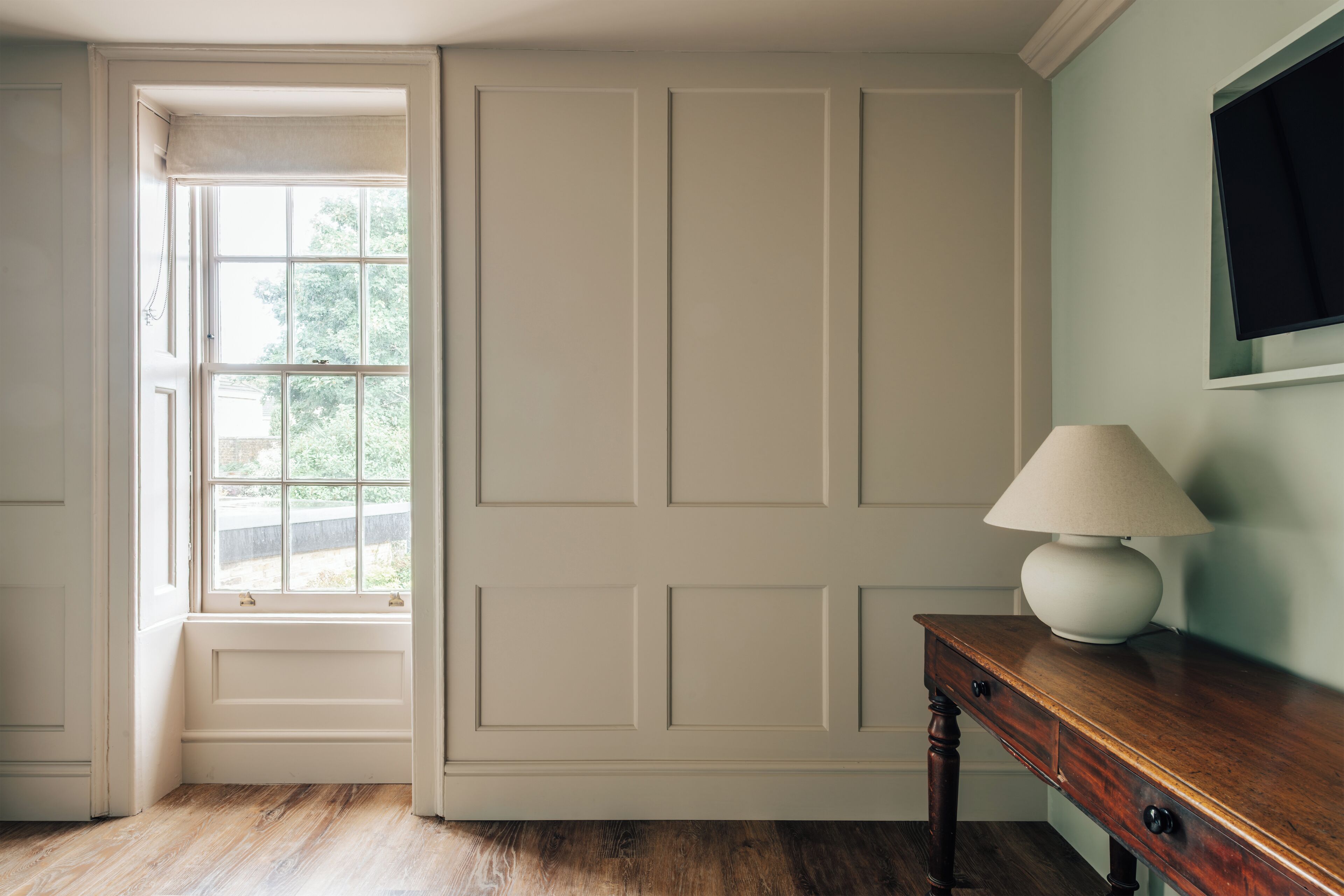 Una habitación con piso de madera, una puerta blanca, un ventanal con vista a follaje y un escritorio de madera con una lámpara.