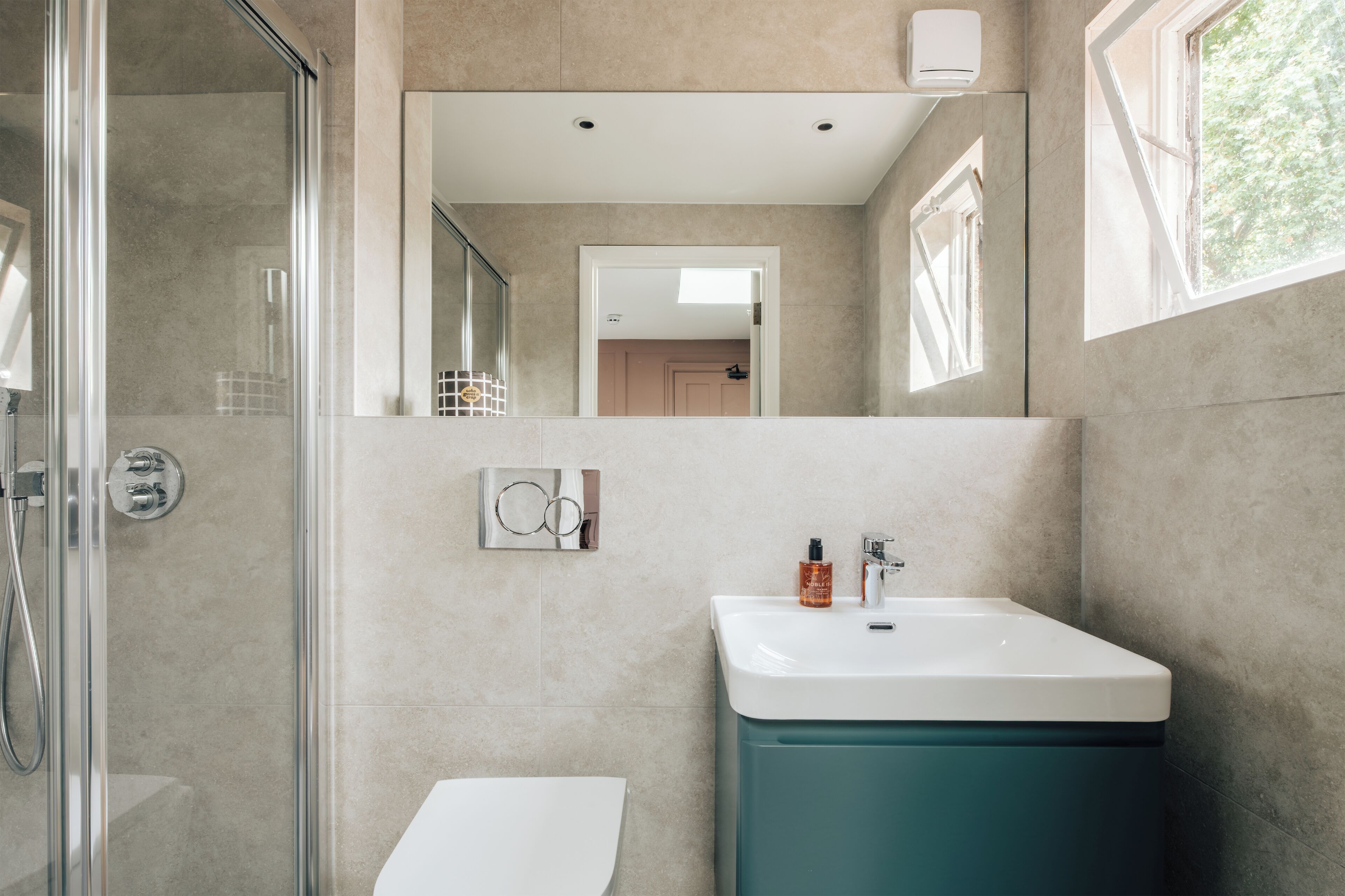 Un baño moderno con cabina de ducha de vidrio, inodoro blanco, tocador verde azulado con lavabo blanco y espejo sobre el lavabo.