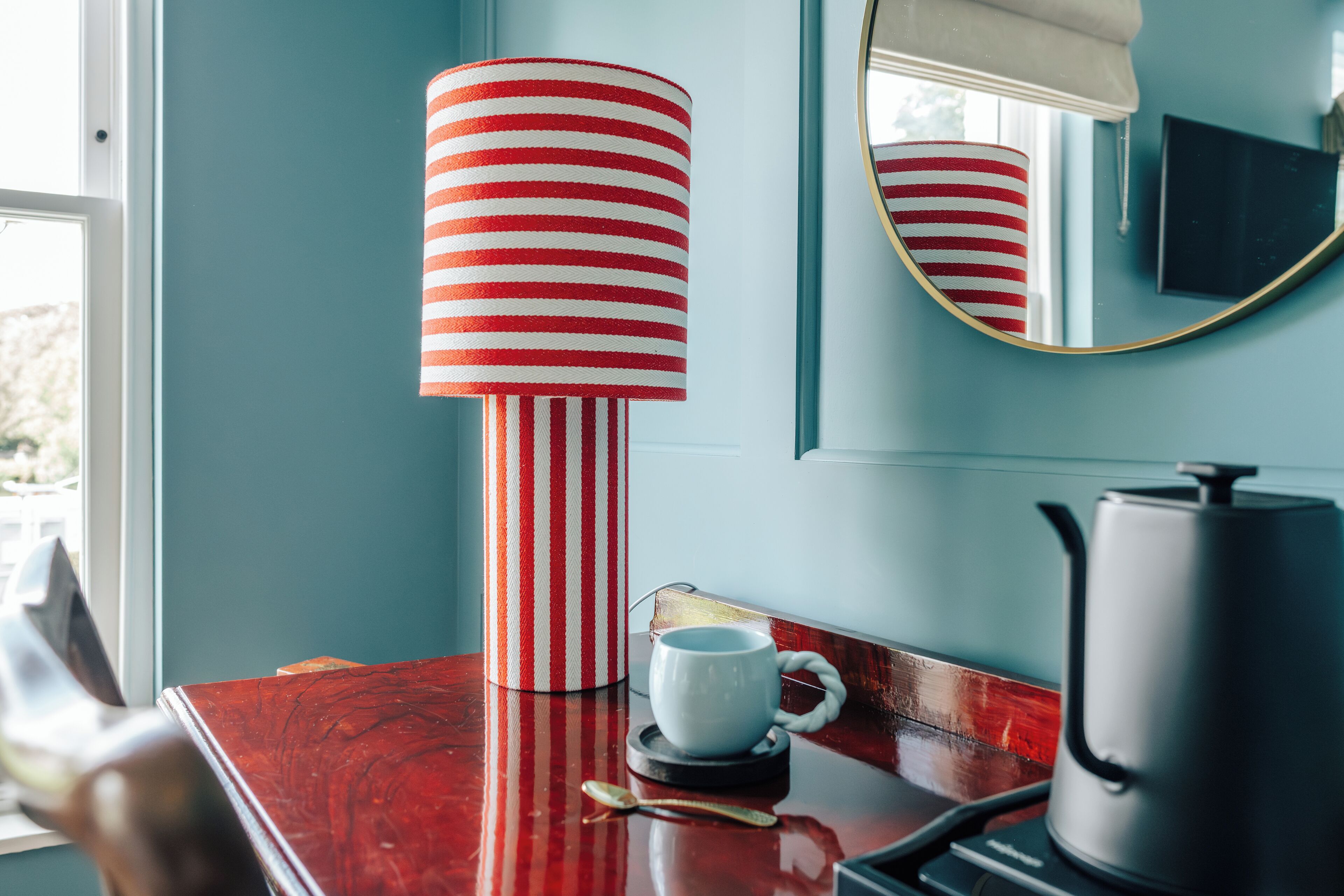 A room with a striped lamp, a white mug, a book, and a kettle on a wooden table.