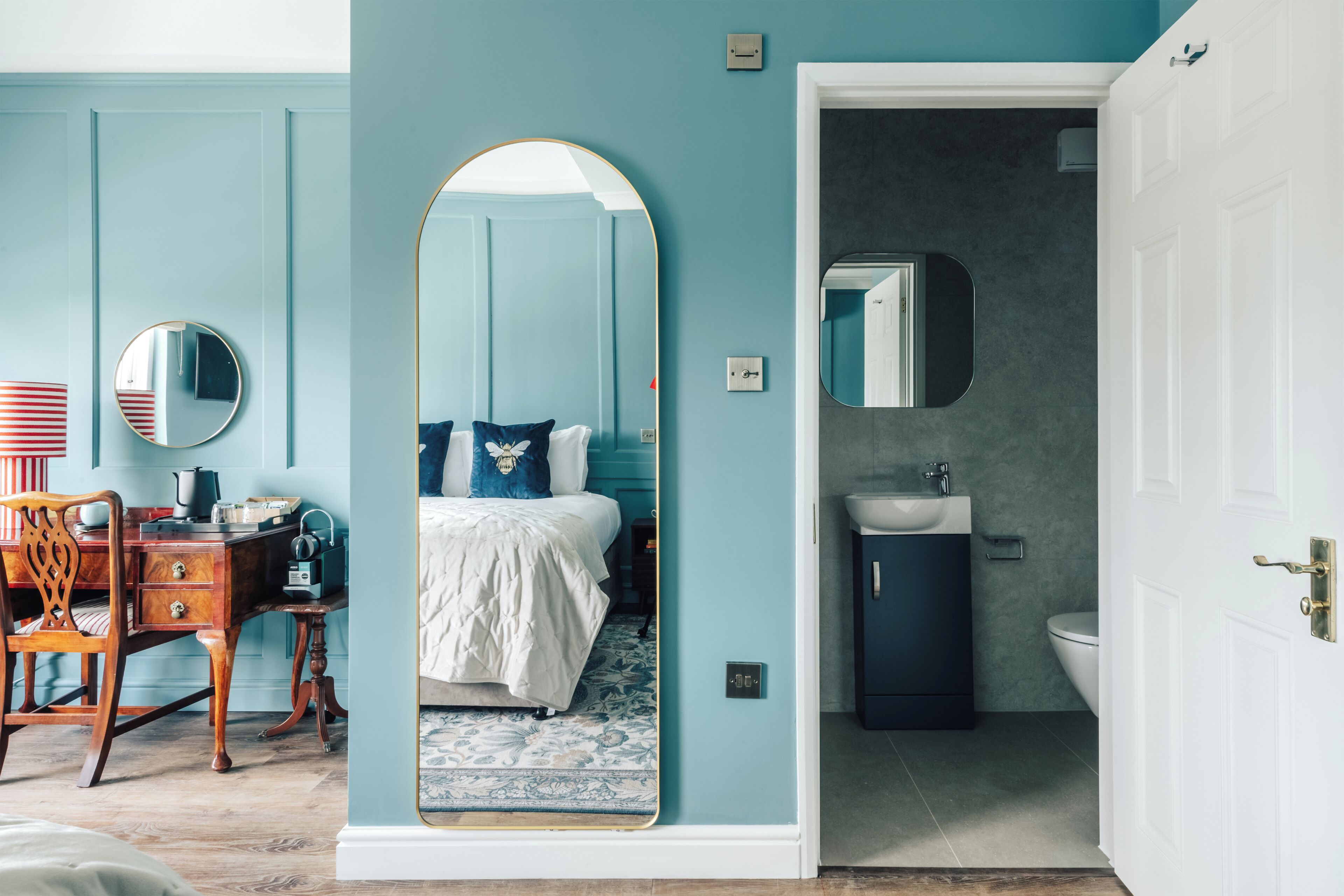 Une chambre à coucher avec un lit, un bureau et une chaise. On peut voir une salle de bain avec un lavabo et des toilettes à travers une porte ouverte.