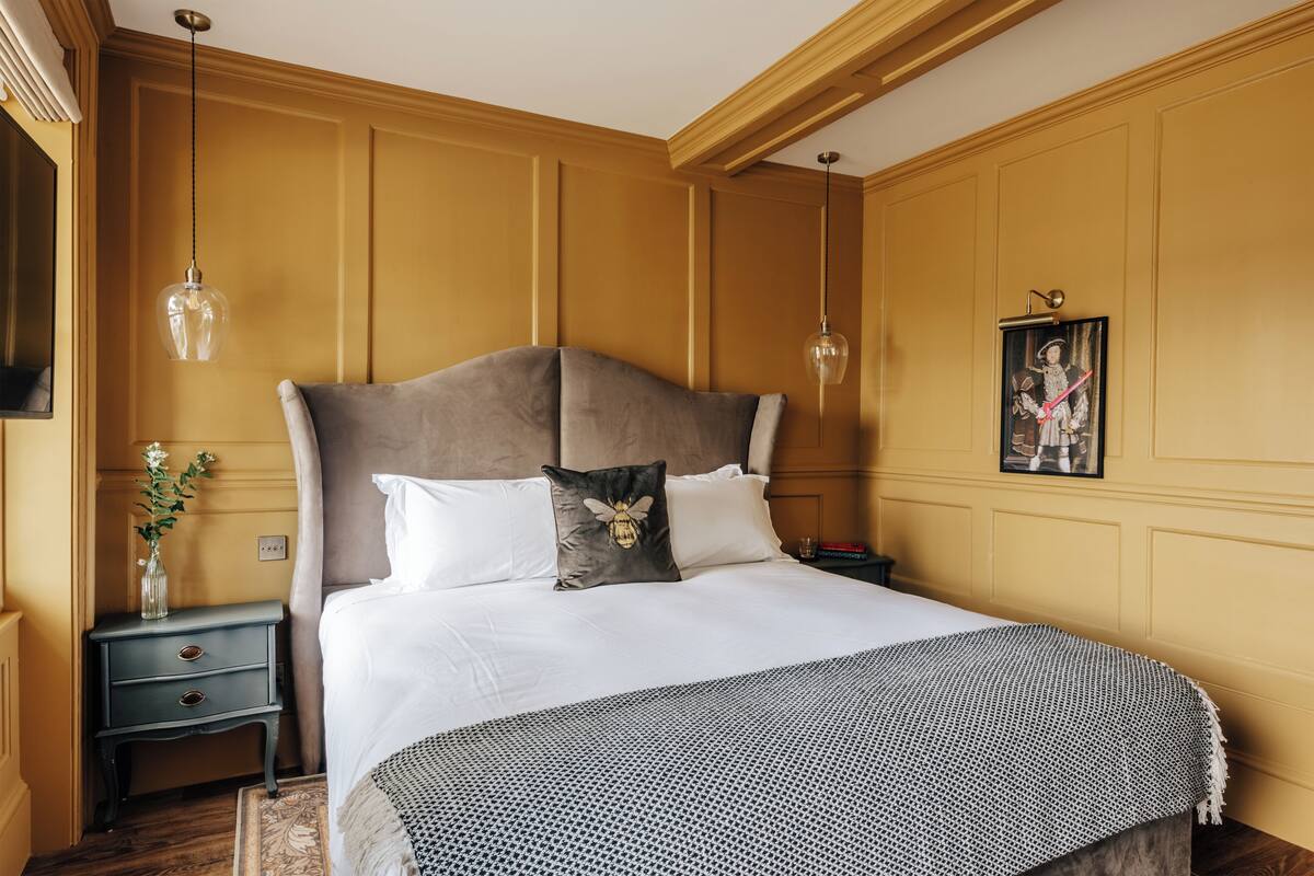 Un lit bien fait, avec un grand oreiller et un coussin décoratif, une table de chevet ornée d’un vase de fleurs et un tableau encadré accroché au mur.