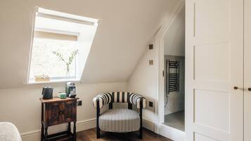 Una habitación acogedora con techo inclinado, una ventana que da a un muro de ladrillo, una silla a rayas, una pequeña mesa de madera con una tetera y un recipiente verde, y una puerta blanca.