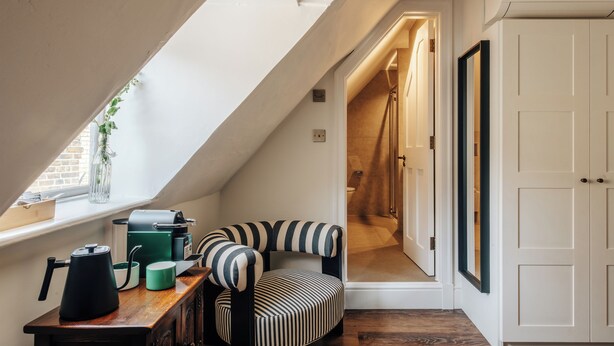A cozy attic room with a sloped ceiling, a striped armchair, a wooden table with a kettle, and a white wardrobe. - Hermes Hotel by Nestor (Kingston Upon Thames)