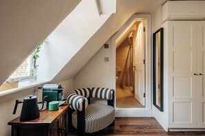 Een gezellig zolderkamertje met een schuine hemel, een gestreept fauteuil, een houten tafel met een waterkoker en een witte garderobe.