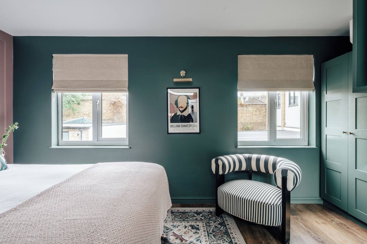Une chambre à coucher comprenant un lit, une chaise, un tableau encadré et deux fenêtres équipées de stores.