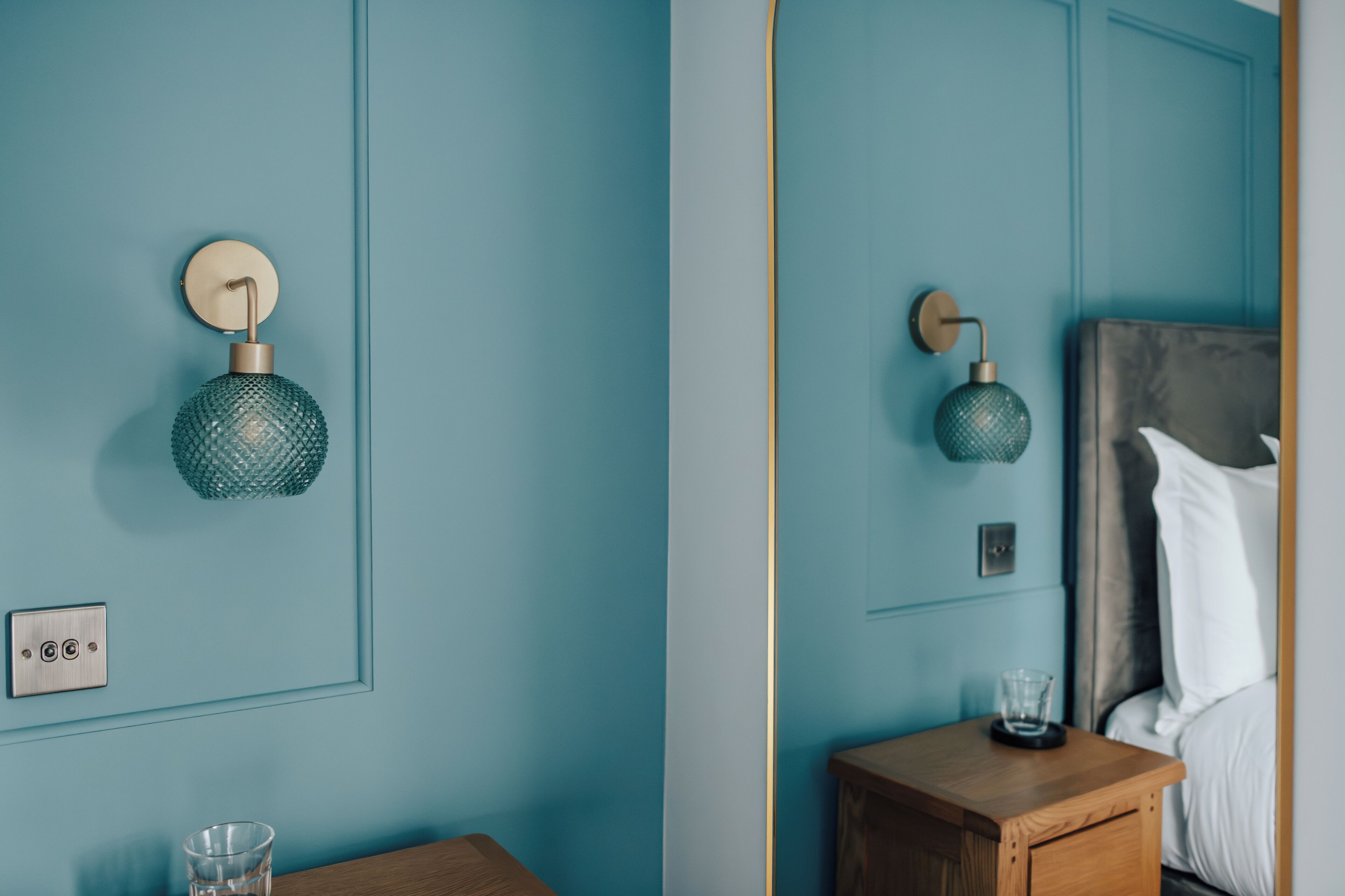 A room with teal walls, a wooden nightstand, a bed with white bedding, and two wall-mounted spherical lamps.
