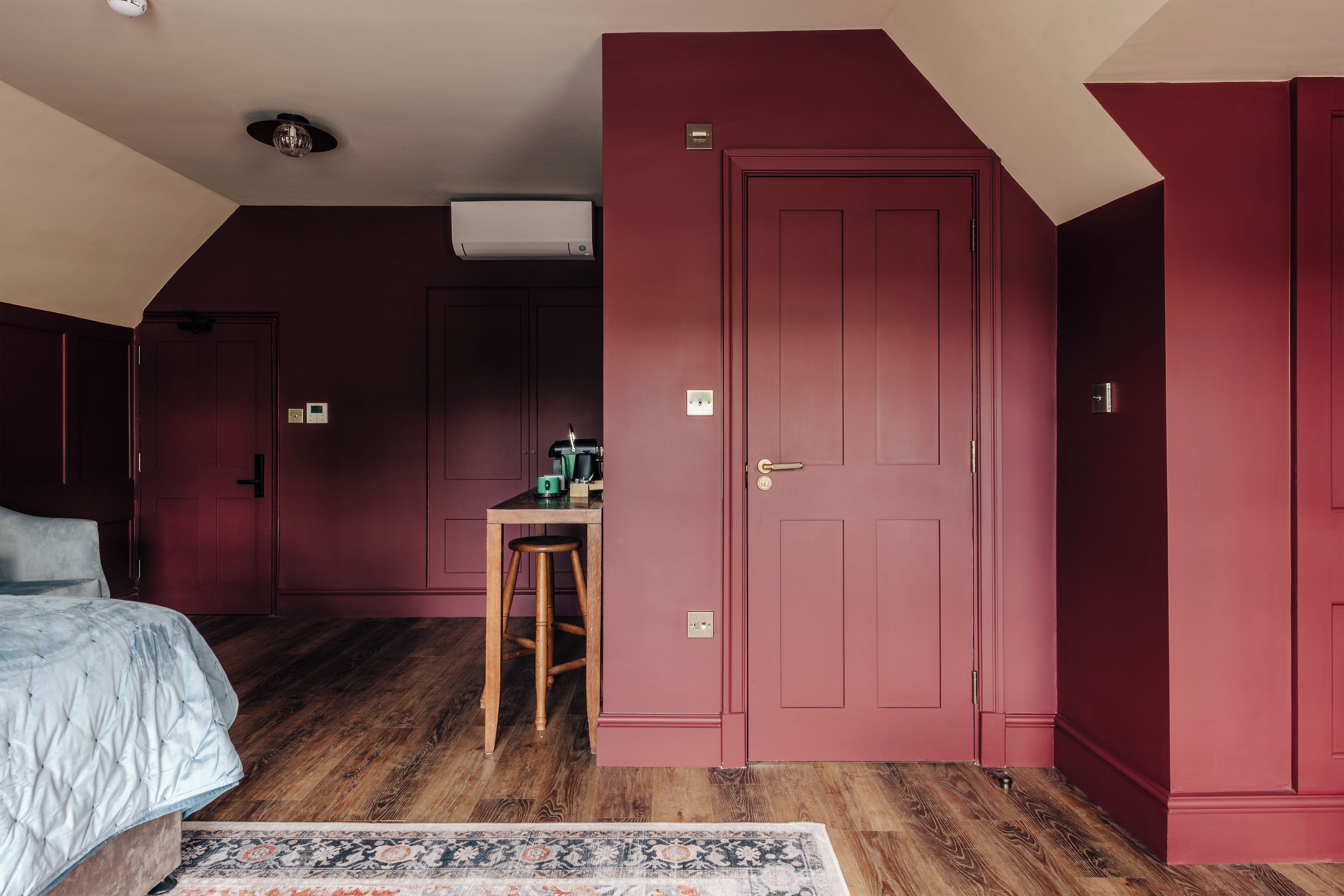 A bedroom with a large bed, a wooden floor, a red door, and a small table with a chair.