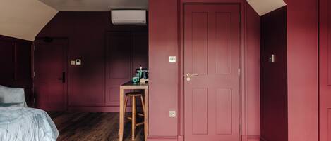 A bedroom with a large bed, a wooden floor, a red door, and a small table with a chair.