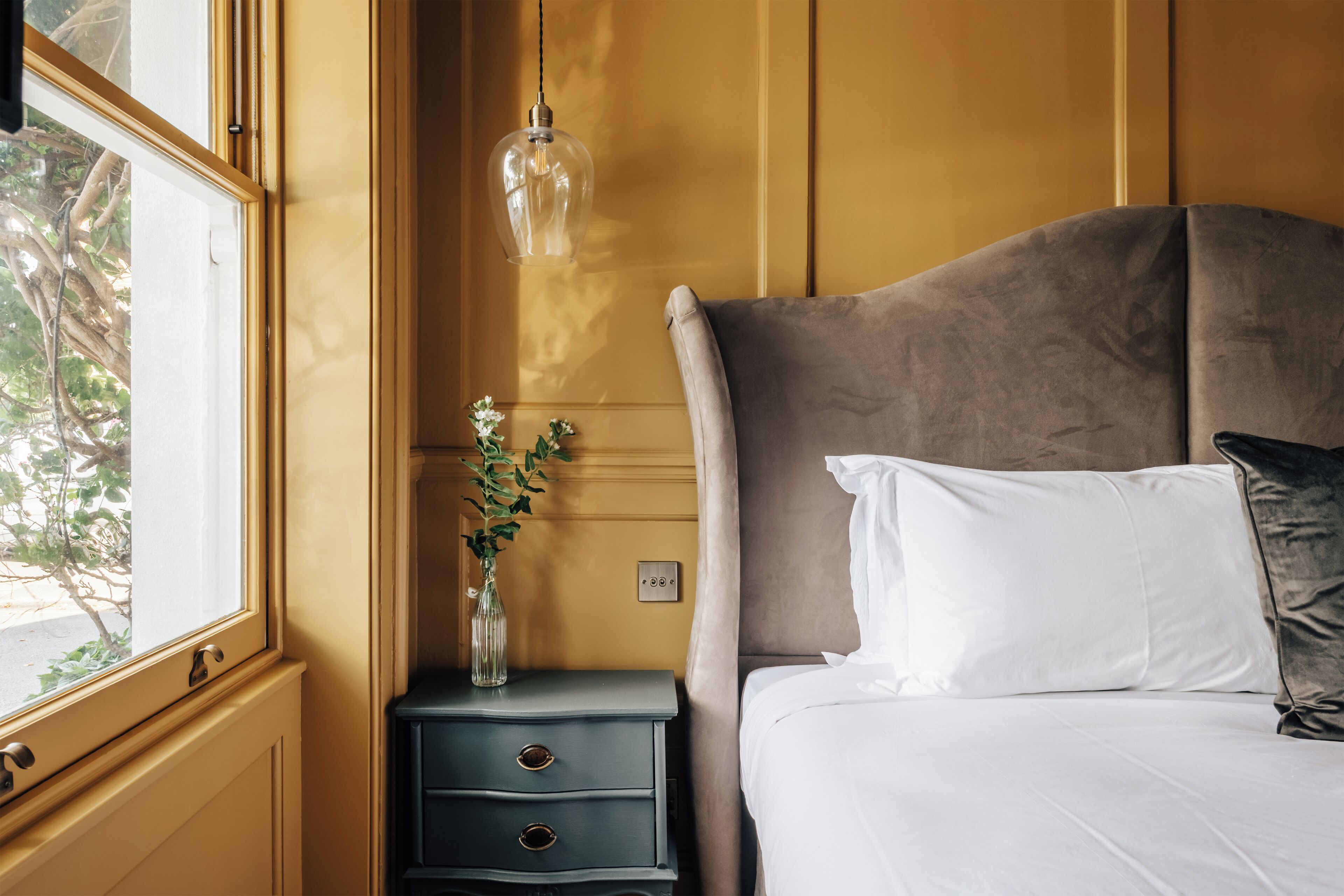 Camera da letto con un grande letto, un comodino con cassetti, un vaso con fiori e una lampada a sospensione.
