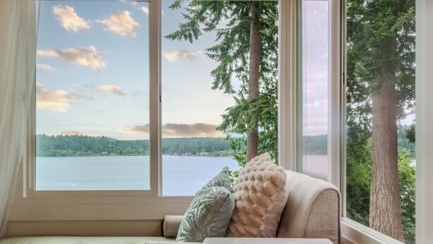 A room with a large window offering a view of a lake and forest. There is a sofa with cushions and a small table. - Oceanfront boho cottage in Bremerton (Bremerton)
