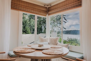 A room with a round table, multiple windows, and a view of a lake and forest. - Oceanfront boho cottage in Bremerton (Bremerton)