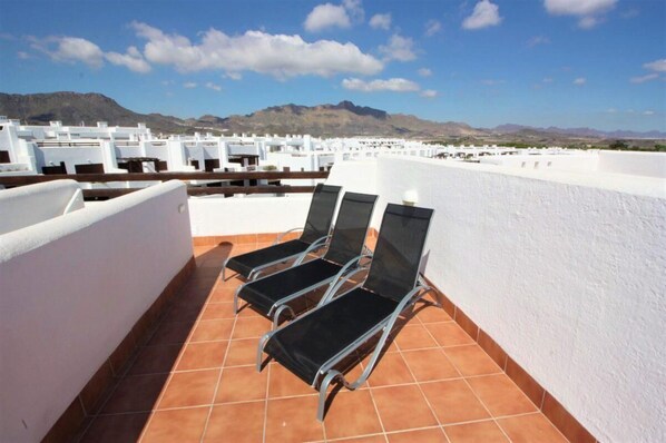 A rooftop terrace with four black lounge chairs and a white wall, overlooking a mountainous landscape.
