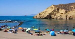 A beach with people sunbathing and swimming, colorful umbrellas, and a rocky cliff in the background.