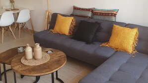 Een woonkamer met een grijze bank, twee gele kussens, een wilgentenen hanglamp, een ronde salontafel met twee keramieken vazen, en een lijst met een afbeelding aan de muur.