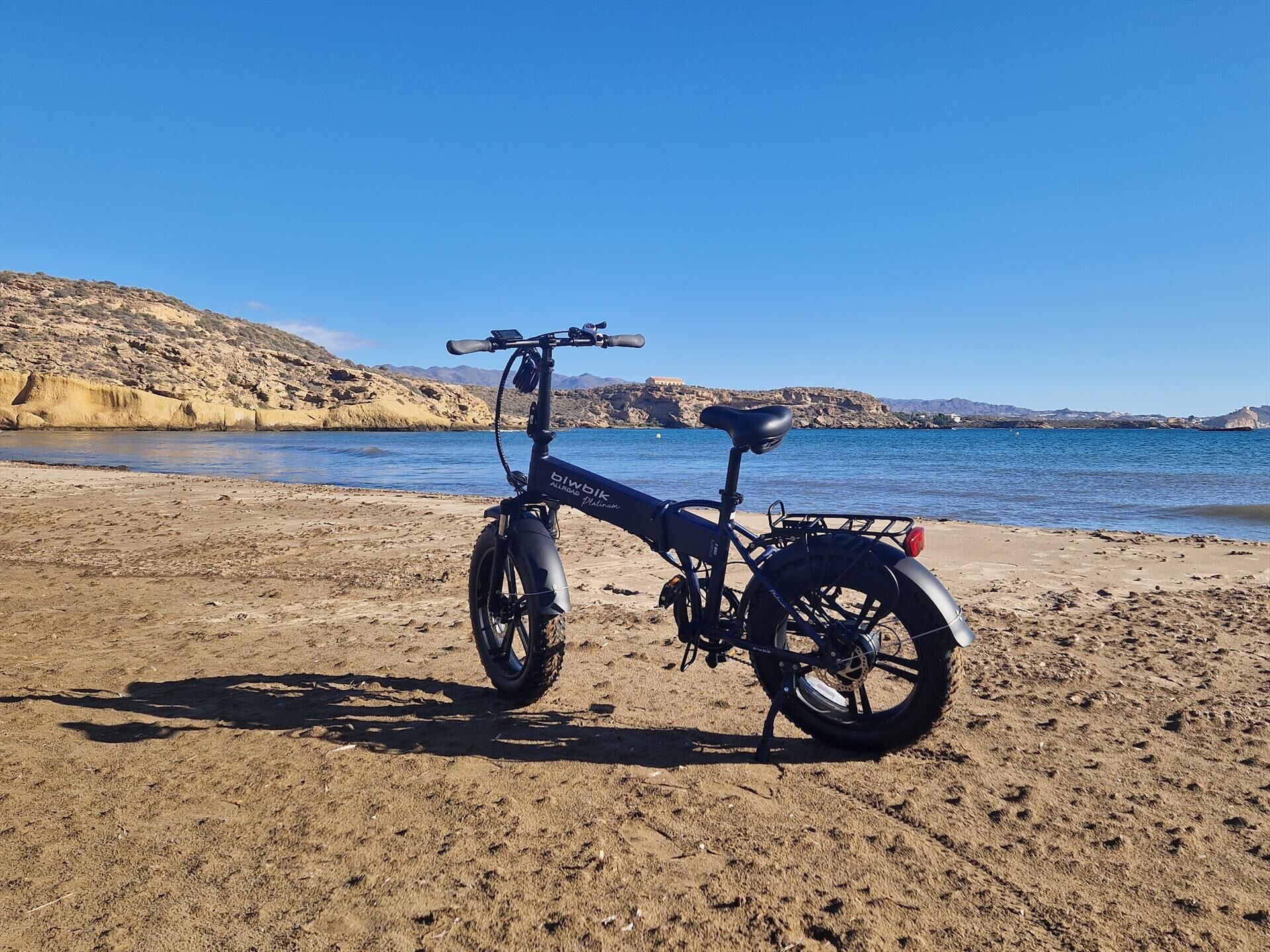 Een fiets geparkeerd op een zandstrand, met een helder uitzicht op de zee en rotsachtige heuvels op de achtergrond.