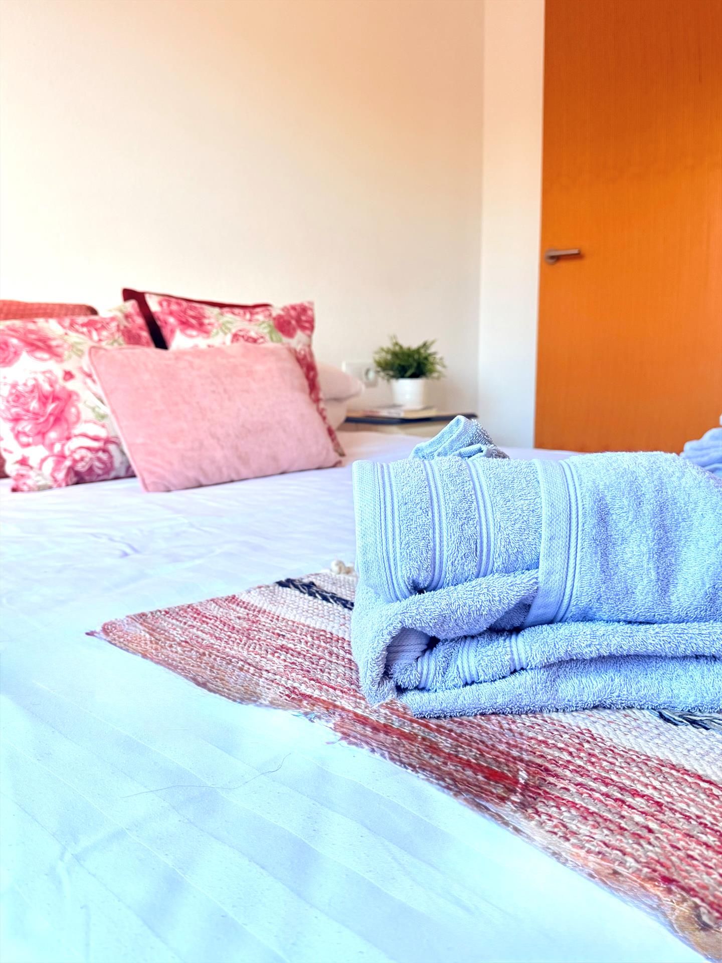 Una cama bien hecha con una toalla azul, un cojín estampado en rojo y blanco, y un cojín con motivos florales.
