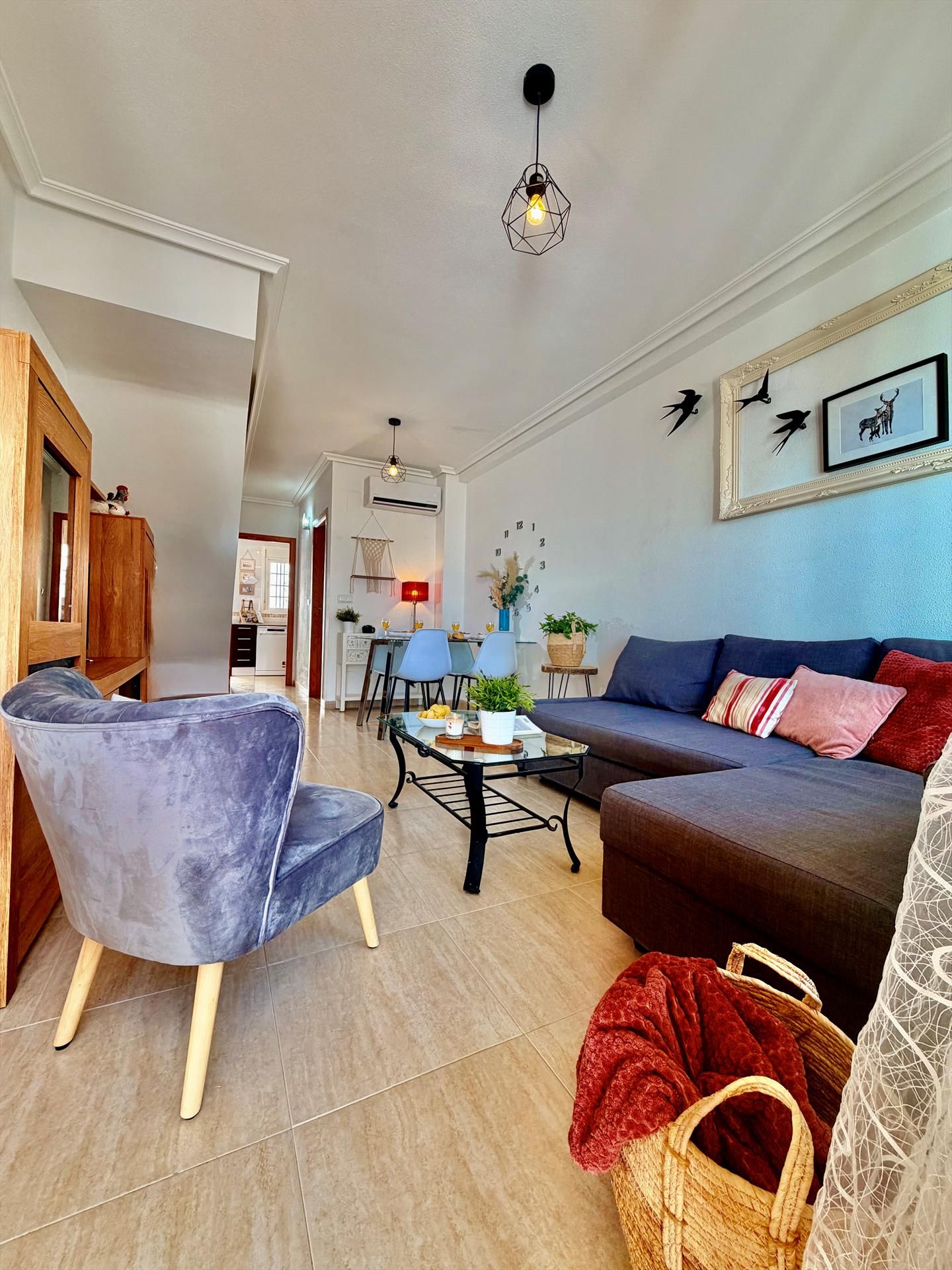 Een moderne woonkamer met een bank, fauteuil en salontafel. Er is een eethoek met stoelen en een tafel. De kamer heeft een lichtgekleurde vloer en witte muren.
