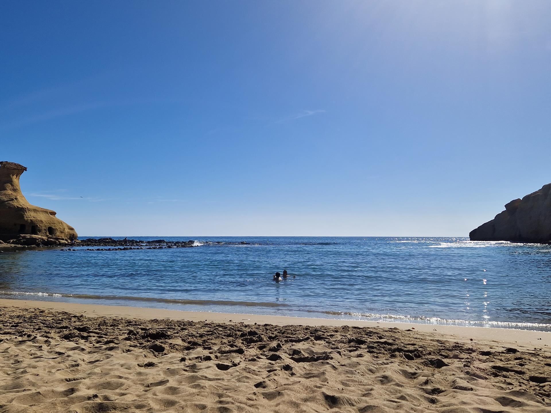 En strand med kristallklart blått vatten, en sandstrand och klippiga branter.