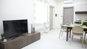 A modern living room with a flat-screen TV on a wooden console, a dining area with a table set for two, and a kitchen with a stove and cabinets.