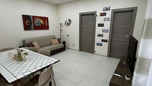 A modern living room with a sofa, a dining table with chairs, a TV, and a wall with photos.