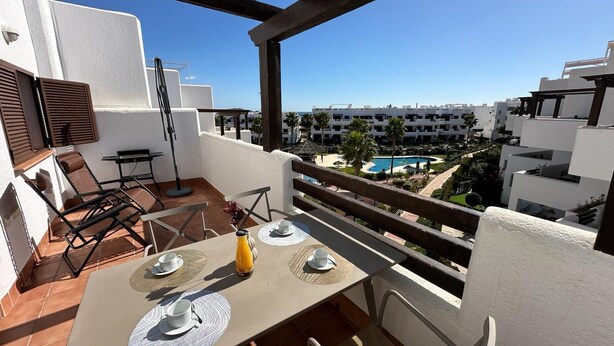 A balcony with a table set for two, overlooking a pool and residential buildings. - Casa Pedro - Two Bedroom Apartment, Sleeps 4 (San Juan de los Terreros)