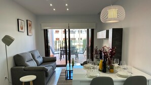 Una sala de estar moderna con un sofá gris, una mesa de comedor lista para cuatro personas y un amplio ventanal con vistas a un patio.